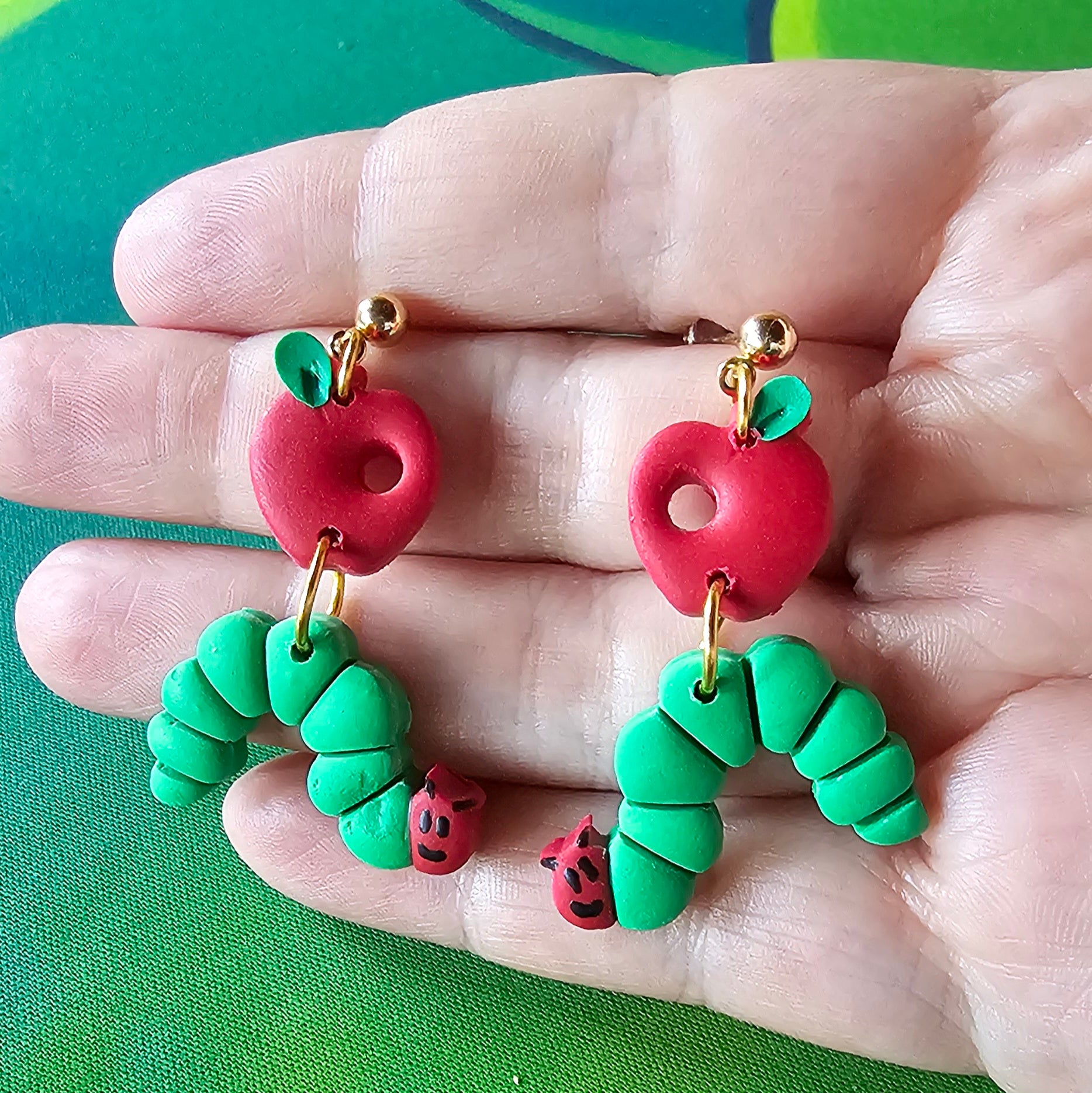 Hungry caterpillar dangle earrings with gold hardware. There is an apple with a hole in it and below dangles the hungry caterpillar. The earrings are held by a hand.